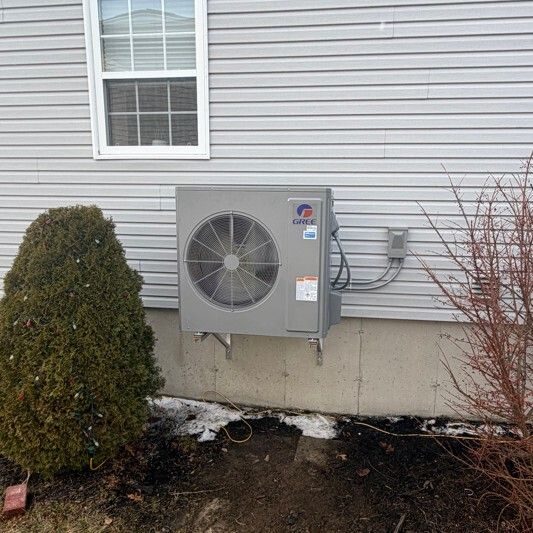 Gree outdoor heat pump unit on a wall bracket beside grey siding in early spring snow